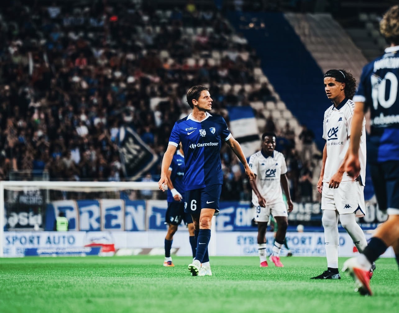 Match du GF38 au Stade des Alpes à Grenoble avec les joueurs sur la pelouse et les tribunes en arrière-plan.