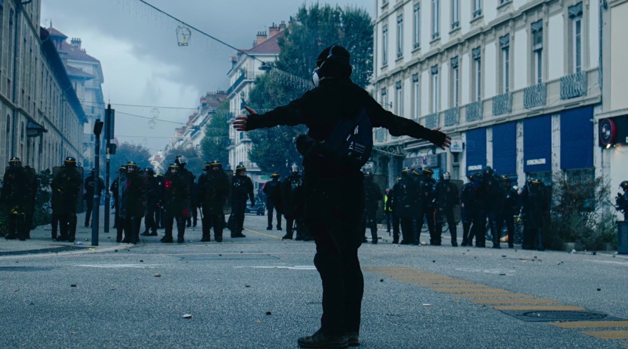 Manifestant les bras ouverts face à des policiers équipés pour le maintien de l'ordre lors des manifestations du 17 octobre 2026 à Grenoble. Une scène diffusée par Chicagre et devenue virale sur X.